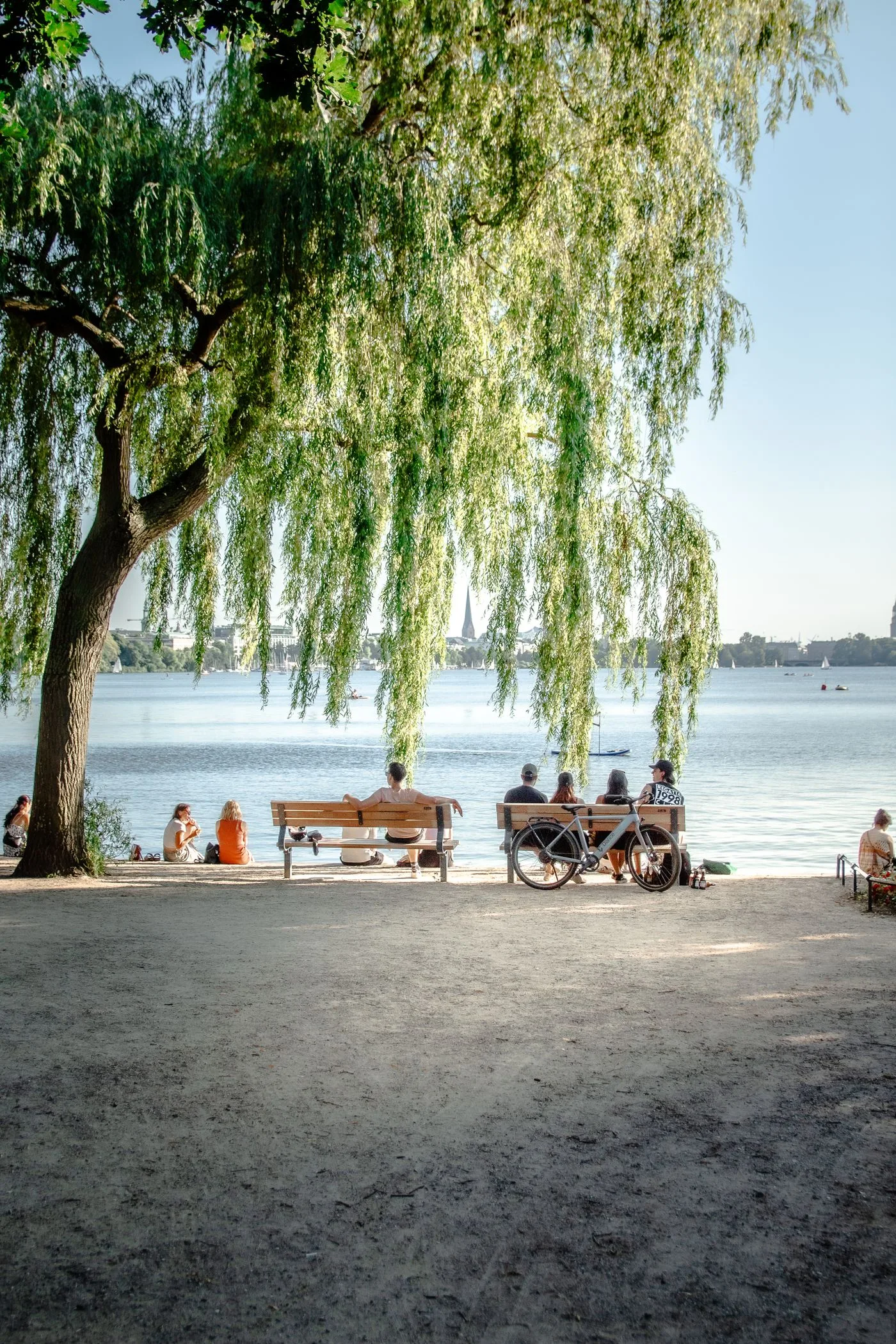 Menschen sitzen auf Bänken unter einer Trauerweide am Alsterufer in Hamburg